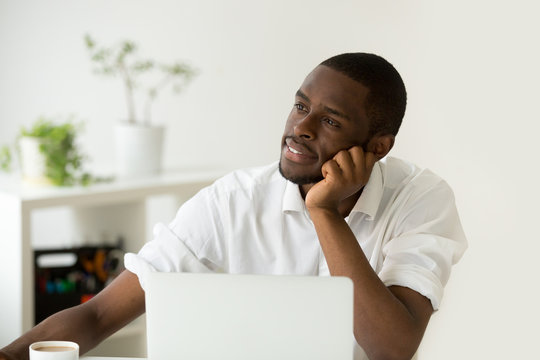 Dreamy african american young man thinking of new idea at workplace, black lazy worker avoiding work lost in thoughts, absent-minded intern dreaming of successful business and happy future career