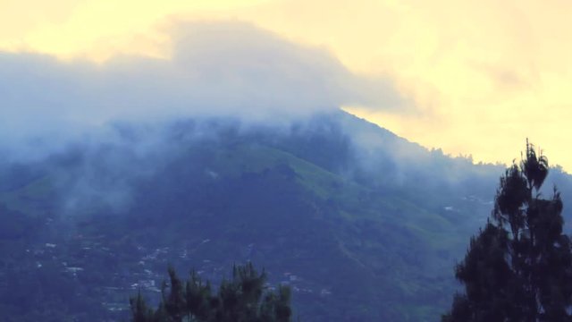 Footage of South American Andes mountain range peak covered by dense fog moving across landscape and village sitting below. Framed by pine trees moving with the wind on the foreground.