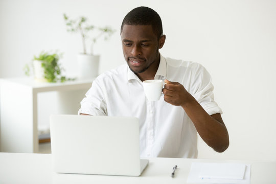 Smiling attractive african american man enjoying coffee while using laptop sitting at home office desk, black businessman checking online news or computer emails relaxing having morning break at work
