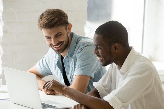 Diverse smiling businessmen watching funny video using laptop together, african and caucasian coworkers having fun talking looking at computer screen, multiracial office workers enjoy online teamwork