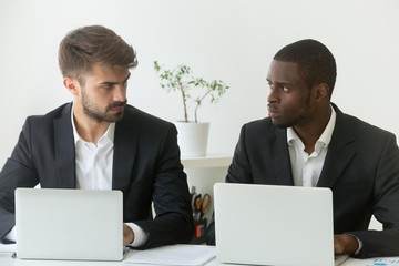 Multiracial office rivals looking at each other with hate envy sitting with laptops, corporate...