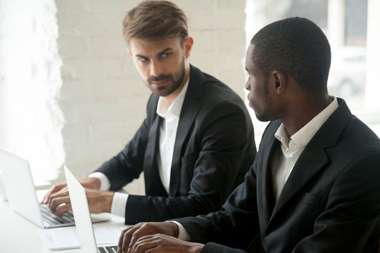 Caucasian businessman looking at african business rival with hate dislike envy, diverse competitors in suits working in office on laptops, multiracial colleagues enemies showing rivalry at work