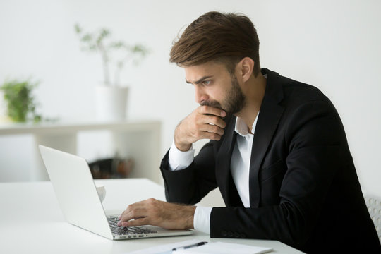 Focused serious businessman in suit thinking reading online news or solving business problem working on laptop looking at screen, worried puzzled executive managing stock market risks using computer