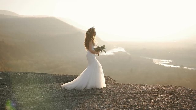 The beautiful girl is walking in the charming modern white dress with wreath of flowers in the mountains in sunset