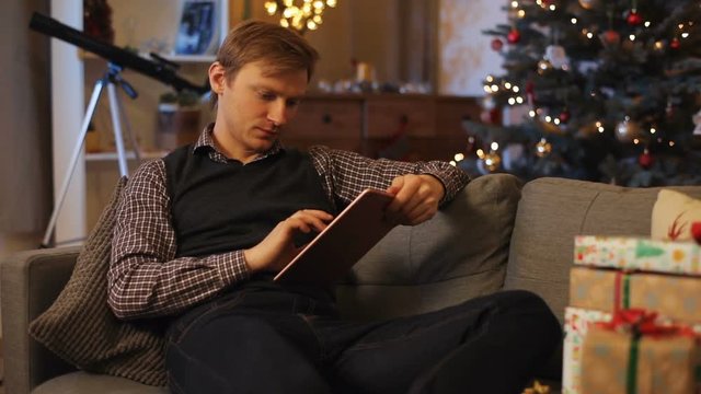 Attractive Caucasian man in vest lying on couch on background of fascinated Christmas tree with decorations. Guy counting gifts, using tablet. Indoor.