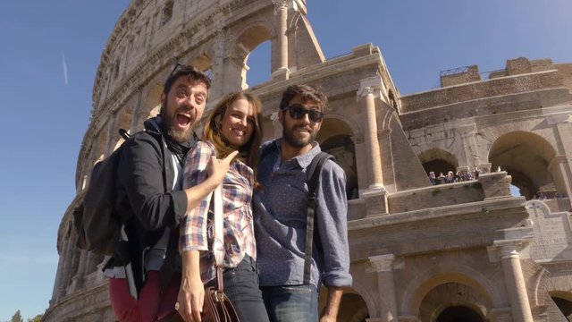 Three young friends tourists standing on pedestal in front of colosseum in rome taking funny hilarious pictures posing with backpacks sunglasses happy beautiful girl long hair slow motion