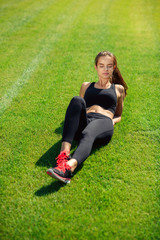 A beautiful sports girl in black sportswear and sneakers resting on green grass after intensive workout.