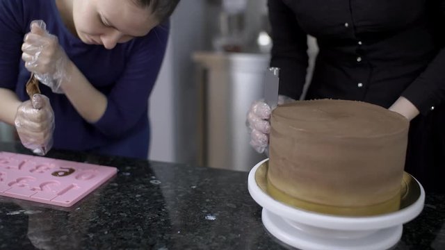 Two female confectioners are working on cake for celebration. Professional is turning the baked candy aroung and deleting pieces with spatula and her colleague is fulfilling pink plastic form with