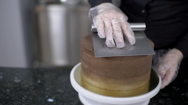 Young professional is putting mastic on surface of cake in kitchen indoors. Female chef is leveling top of chocolate pie on table in pastry shop. Talented woman is working on delicious cake using