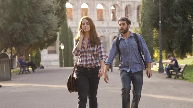 Beautiful girl walks in park road talking on phone and attractive young man boyfriend behind her taps shoulder run funny prank trees colosseum in background in rome at sunset lovely beautiful woman