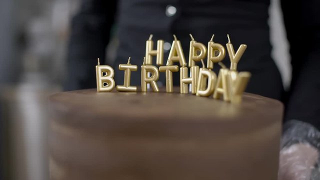 Delicious cake is being observed at the kitchen of confectionery shop. Professional in black shirt and white gloves is turing the baked chocolate candy around and Happy birthday is witten on the top