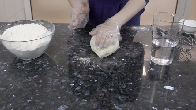 Young woman is kneading dough on table in kitchen at home. Female kneads elastic mass with hands on desk in cuisine indoors. Experienced housewife in gloves prepares wheat dough for pie, touching it
