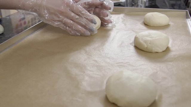 Female baker is putting small pies to baking sheet, close up. Dish is covered with brown paper and pieces of dough are lying in a row. Woman with beautiful manicure is wearing gloves to roll the round