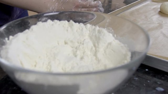 Woman is putting pies to the baking sheet, close up. The dish, covered with brown paper is lying on black table, near glass bowl with powder and female baker in gloves is forming rounds from dough