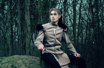 young man in the costume of a barbarian sitting on a stone in the dark forest