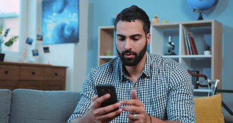 Close up attractive young man typing message on the phone smiling feel happy sit on sofa home portrait smartphone hands electronic app use hold connection leisure lifestyle communication touching