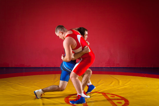 Two strong wrestlers in blue and red wrestling tights are wrestlng and doing grapple on a yellow wrestling carpet in the gym.