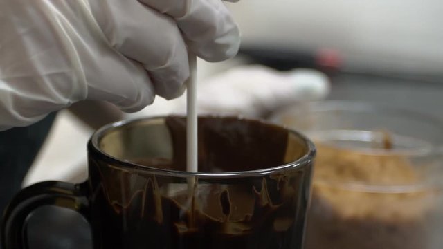 Close up of lollypop is being covered with hot chocolate. Confectioner in white gloves is putting the dark candy into the cup with sweet pastry, and lift it up and the droppings are falling. Glass
