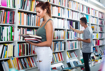 portrait of  teenage girl customer looking at open book standing among bookshelves