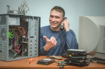 Happy cheerful computer technician engineer of user support service is holding in hands a phone and is showing on broken PC.