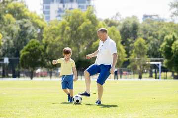 young happy father and excited little 7 or 8 years old son playing together soccer football on city park garden running on grass kicking the ball