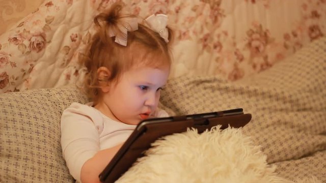 Child Looks cartoons and plays downloaded application on a smart phone close-up. A little cute funny girl lies in bed under the blanket and looks at the white phone screen.