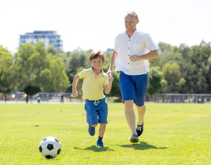 Obraz premium young happy father and excited little 7 or 8 years old son playing together soccer football on city park garden running on grass kicking the ball