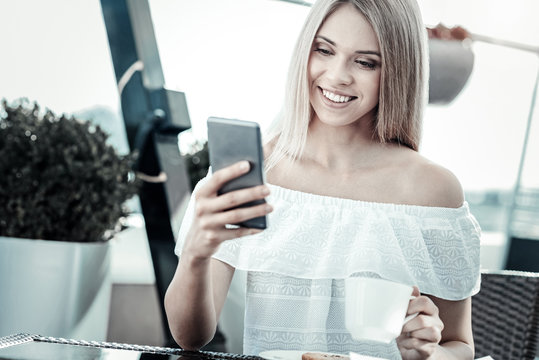 Modern generation. Cheerful nice young woman smiling and looking at her smartphone screen while using it