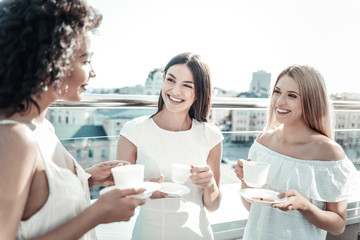 Pleasurable meeting. Delighted positive pleasant women standing on the rooftop and having coffee while meeting at the restaurant