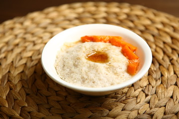 Low-calorie porridge for a healthy diet and lifestyle. Barley with honey and bits of orange pumpkin in a deep white plate. Dark background, wooden table and wicker stand