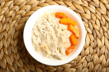 Low-calorie porridge for a healthy diet and lifestyle. Barley with honey and bits of orange pumpkin in a deep white plate. Dark background, wooden table and wicker stand