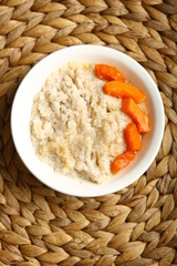 Low-calorie porridge for a healthy diet and lifestyle. Barley with honey and bits of orange pumpkin in a deep white plate. Dark background, wooden table and wicker stand