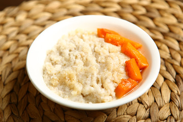 Low-calorie porridge for a healthy diet and lifestyle. Barley with honey and bits of orange pumpkin in a deep white plate. Dark background, wooden table and wicker stand