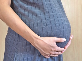 Close-up of Asian pregnant woman in maternity clothes holds hands on belly on wooden background. Image of pregnancy and maternity concept.