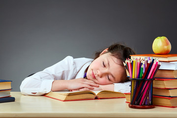 Sleepy and tired beautiful cute little girl leaning on thick books on the grey background