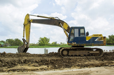 Obraz premium Old Backhoe is working dig ground. Dam construction along the water barrier. At riverside nature in countryside. Has a background landscape river and sky.