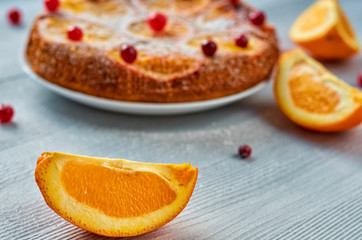 Fototapeta na wymiar Juicy orange slice on the foreground close up with free copy space. Homemade orange cake with fresh red cranberries and oranges on the blurred background. Citrus cake on the gray table. Side view