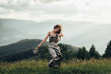 Naklejka na ściany i meble A beautiful girl in a dress and a bouquet of flowers in her hands, on a green glade dancing among the peaks of the mountains