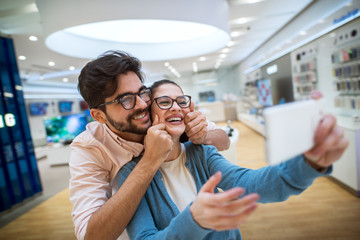 Lovely cheerful hipster couple in the tech store taking a selfie with the shop phone.