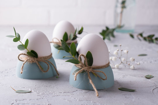 Easter  festive table setting with white chicken  eggs in eggs cups, leaf sprigs of eucalyptus. On a gray concrete background.