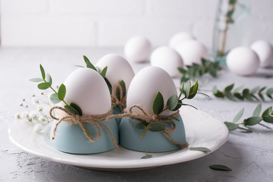 Easter  festive table setting with white chicken  eggs in eggs cups, leaf sprigs of eucalyptus. On a gray concrete background.
