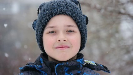 A positive closeup of a small boy in a knitted hat with balls and a blue parka. He stands and smiles in a snowy alley in winter. 