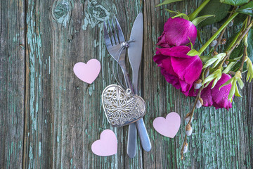 Festive table place setting for romantic dinner or valentine's day with bouquet of purple roses, cutlery, hearts on rustic wooden background with copy space