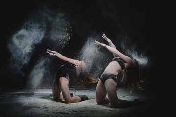 Two girls dance with flour in studio on black background, lights behind them and people helped girls