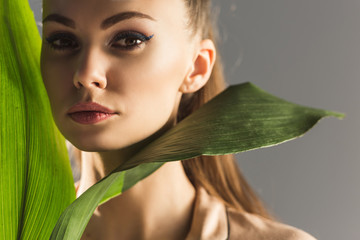 attractive trendy girl with green leaves looking at camera, isolated on grey