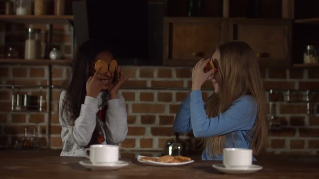 Smiling multiracial little girls covering their eyes with cookies during breakfast while sitting in moden kitchen. Cute playful kids with toothy smile fooling around, hiding their eyes behind cookies.