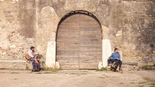 Happy young couple backpackers tourists sitting near wooden doorway of medieval building in parco degli acquedotti park ruins in rome at sunrise with guitar and map guide slow motion steadycam