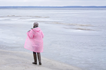 Young woman walking on beach watching the frozen water in a pink rain coat
