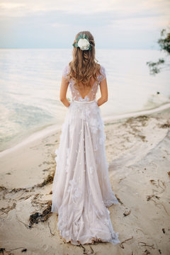 Bride walks on river bank with flowers in hair, photo from back