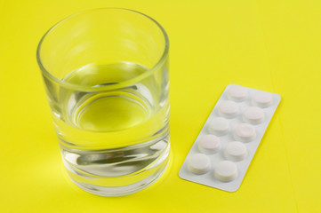 Pills in white blister pack beside water on transparent glass in background of yellow paper